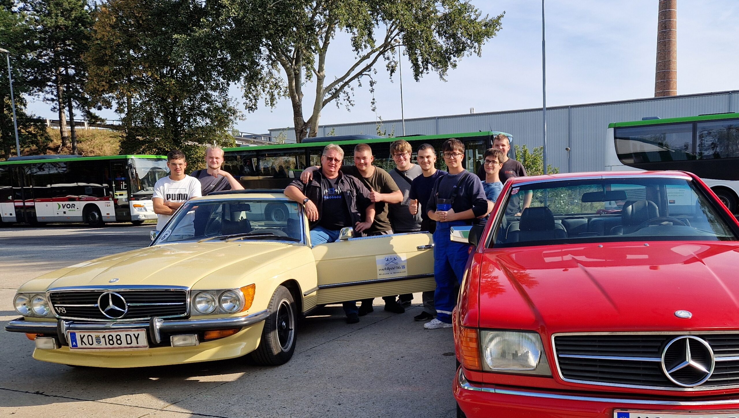 Gruppe von Männern steht neben gelbem Mercedes-Oldtimer und roten Mercedes vor mehreren Bussen.